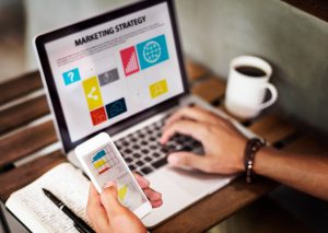 person typing on laptop that displays the title 'Marketing Strategy', and holding a smartphone in his other hand