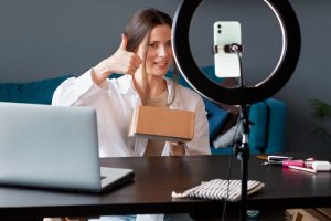 A girl taking a selfie video with her phone and a ring light, sitting in front of her laptop