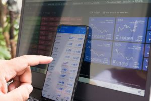 financial-apps-business - One Click Root Person clicking on a financial app on their smartphone, with a similar app showing on a monitor behind the phone